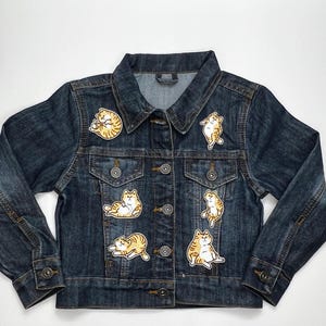 May include: A dark wash denim jacket decorated with several patches of cartoon cats in different poses. The patches depict orange and white tabby cats. The jacket has a button-up front, two chest pockets, and long sleeves.