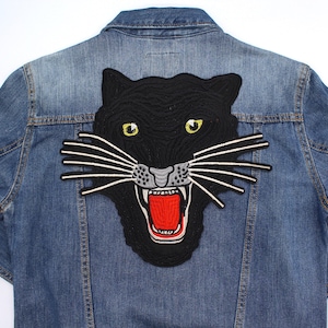 May include: A light blue denim jacket featuring a large embroidered black panther patch. The panther has yellow eyes, white whiskers, and a red open mouth. The jacket is a classic style.
