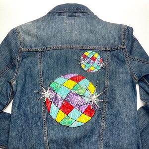 May include: A blue denim jacket featuring two sequined patches on the back. The patches have a mosaic design in red, yellow, blue, and purple sequins. Silver starburst embellishments are also present.