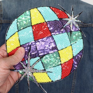 May include: A large, round sequined patch with a colourful geometric design. The patch features squares of yellow, red, purple, and teal sequins. Two starbursts with silver sequins are attached. Shown on a denim jacket.