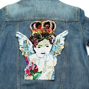 May include: A denim jacket in light blue, adorned with a sequined angel motif on the back. The patch depicts a cherub wearing a crown, with wings and a floral bouquet. The sequins are in gold, red, and blue.