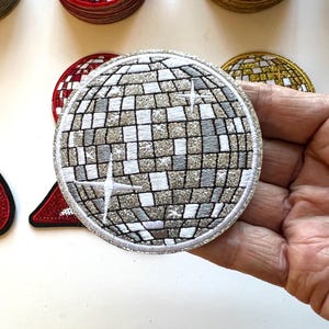 May include: A circular embroidered patch featuring a silver and white disco ball design with a grid of square tiles. The patch has a shimmering, glittery texture throughout the surface and includes white starburst accents across the sphere.