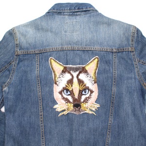 May include: A blue denim jacket with a Siamese cat patch on the back. The patch shows a cat's face with blue eyes, brown and tan fur, and gold detailing. The jacket is a medium wash.