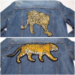 Peut inclure: Deux vestes en jean, chacune avec un grand écusson animalier au dos. L'une présente un écusson léopard avec des paillettes dorées, l'autre un écusson tigre avec broderie jaune et noire.