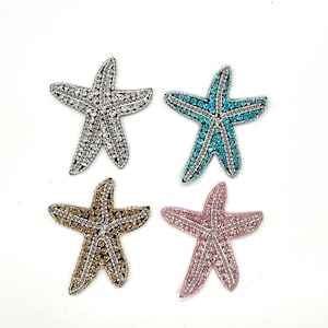 May include: Four bejeweled starfish in silver, turquoise, gold, and pink. Each starfish is embellished with small, sparkling stones, creating a shimmering effect. The starfish are arranged against a white background, highlighting their vibrant colours and intricate details.