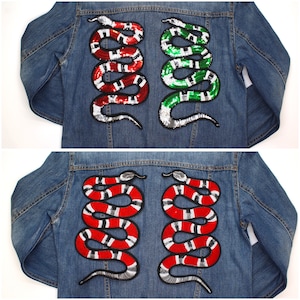 Puede incluir: Dos chaquetas vaqueras con parches de serpientes con lentejuelas. Una chaqueta presenta serpientes rojas y blancas, la otra serpientes verdes y blancas. Las serpientes están enrolladas y tienen contornos negros. Las chaquetas son de un lavado azul medio.