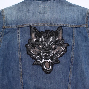 May include: A medium-wash denim jacket with a large embroidered wolf patch. The patch depicts a snarling wolf's head in black, grey, and white. The jacket's back features the patch, adding a bold, decorative element.