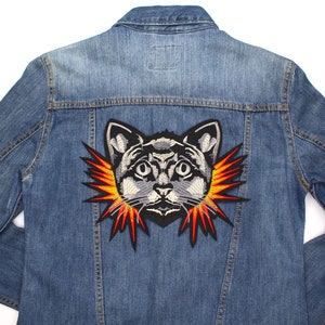 Può includere: Giacca di jeans blu chiaro con una grande toppa ricamata a forma di gatto sul retro. La toppa presenta un volto di gatto dettagliato con un'esplosione di raggi arancioni e gialli. La giacca ha un design classico.