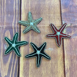 May include: Four starfish-shaped patches decorated with small beads. One patch is gold, one is red, one is dark green, and one is black. Each patch features a central cluster of white beads and white beaded outlines along the five arms.