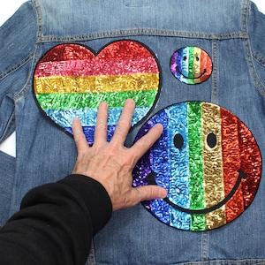 May include: A light-wash denim jacket adorned with three sequined patches. These include a rainbow heart, a rainbow smiley face, and a smaller rainbow smiley face. The sequins shimmer. The jacket is a classic denim style.