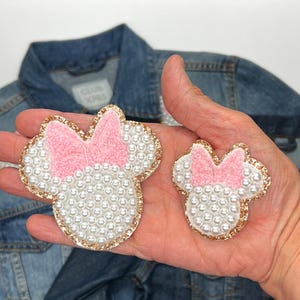 May include: Two Minnie Mouse-shaped patches. The patches have a white pearl-like base, a pink bow, and a gold glitter outline. The patches are in different sizes. A denim jacket is in the background.