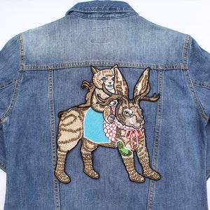 May include: A blue denim jacket with an embroidered patch. The patch depicts a cat riding a rabbit-like creature with antlers. The patch is in brown, tan, blue, and red colours.