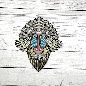 May include: Embroidered patch depicting a stylised mandrill face. The patch features a detailed design with a mix of colours, including blue, yellow, pink, and black. The mandrill's face is the central focus, with intricate patterns and textures.