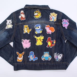 May include: A dark blue denim jacket decorated with colourful embroidered patches of cartoon characters. The patches feature yellow, red, blue, pink, and white hues. The jacket has a classic collar and a button-up front.