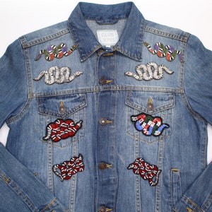 May include: A blue denim jacket adorned with several embroidered snake patches. The patches feature colourful bead and sequin detailing. The jacket has a button closure, two chest pockets, and the text "CLUB XPRS" on the label.
