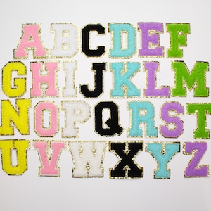 May include: A full alphabet of embroidered fabric letters in various colours, including pink, yellow, green, and blue. Each letter has a gold glitter border. The letters are displayed on a white background.