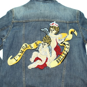 May include: A mid-wash denim jacket with an embroidered cherub patch on the back. The patch includes two cherubs, a crown, and a banner with the words "L'AMORE E BELLEZZA" in yellow. The jacket has classic denim detailing.