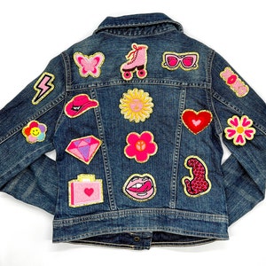 May include: A dark denim jacket decorated with various patches. Patches include a pink butterfly, roller skate, sunglasses, and a flower. Other patches include a diamond, lips, and a heart. The patches are outlined in gold.
