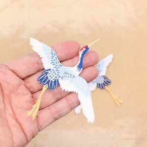 May include: Two embroidered crane patches. The cranes feature white wings with light blue detailing, dark blue tail feathers, and red, blue, and yellow details on their heads and legs. The patches are held in a hand.