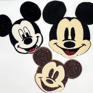 May include: Three Mickey Mouse-shaped decorative items. Two are made with black and cream-coloured yarn, and one is brown with cream-coloured sequins. All three have black ears, cream-coloured faces, and red mouths.