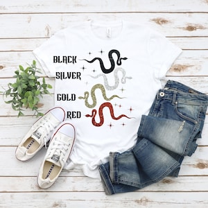 May include: A white t-shirt featuring glittery snake designs in black, silver, gold, and red, with corresponding text. The words "BLACK", "SILVER", "GOLD", and "RED" are printed above each snake. White trainers and blue jeans are also visible.
