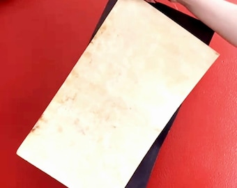 May include: A close-up of a blank, off-white, rectangular piece of paper being held by a hand. The paper is slightly wrinkled and has a brown, aged appearance. The paper is being held over a black surface against a red background.