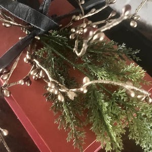 May include: A red gift box tied with a black ribbon, adorned with gold-colored berry branches and green sprigs. The arrangement suggests a festive or decorative theme, possibly for a holiday or special occasion.