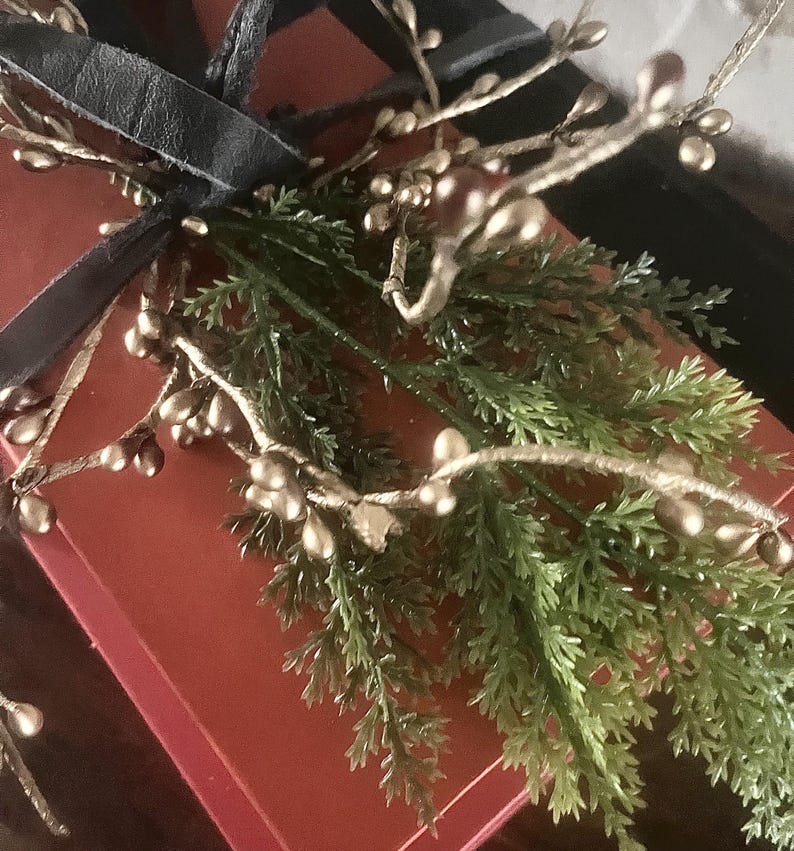 May include: A red gift box tied with a black ribbon, adorned with gold-colored berry branches and green sprigs. The arrangement suggests a festive or decorative theme, possibly for a holiday or special occasion.