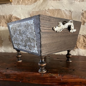 May include: A dark brown wooden box with ornate white handles and decorative accents. The box has four small, turned legs and a distressed, vintage appearance. The front panel features text and a lace-like border.