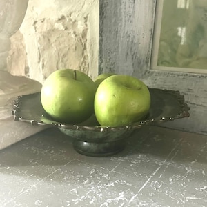 May include: A decorative, antique-style, green-toned metal fruit bowl with a scalloped edge, holding three green apples. The bowl sits on a pedestal base. The background includes a textured wall and a white-framed picture.
