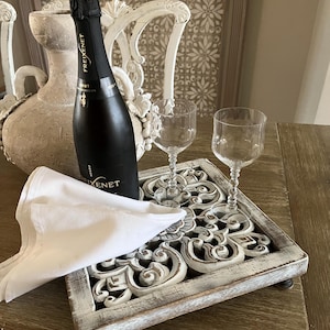 May include: A black bottle of Freixenet sparkling wine, a white napkin, and two clear wine glasses rest on a decorative, distressed white tray with carved details. The tray sits on a wooden table.
