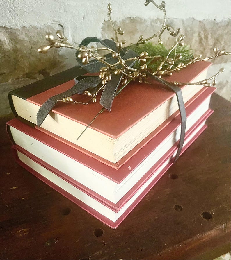 May include: A stack of three books with red covers and white pages, bound together with a dark gray ribbon. The top of the books are decorated with gold-colored sprigs and green foliage, creating a rustic, decorative arrangement.