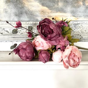 May include: A bouquet of faux peonies in shades of pink and burgundy rests on a white surface. The arrangement includes large, fully bloomed flowers and smaller buds, with green foliage accents. The background features a decorative white frame.