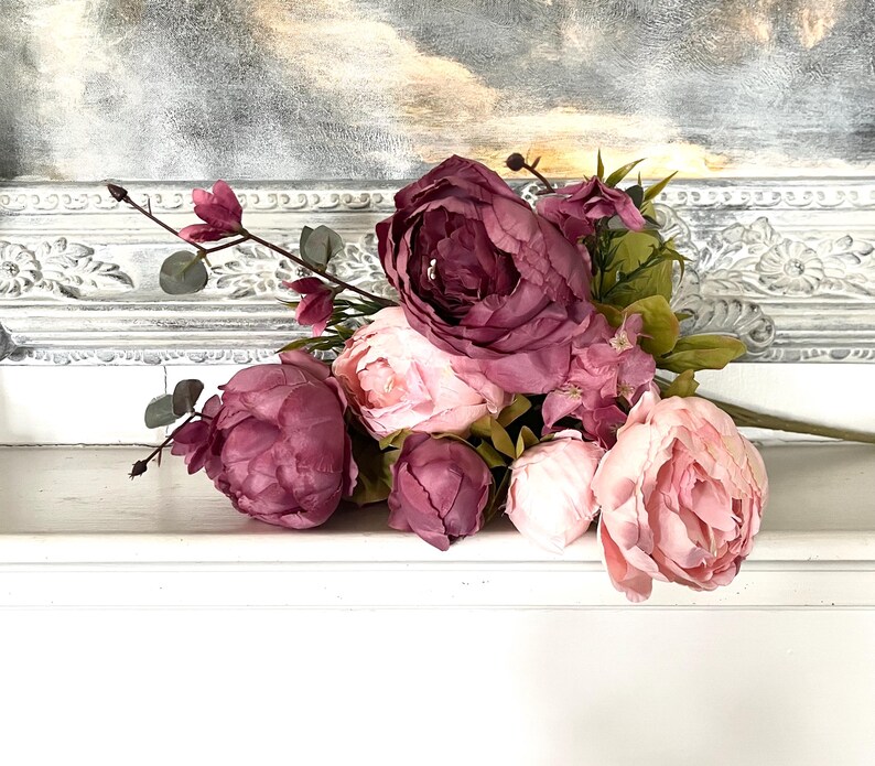 May include: A bouquet of faux peonies in shades of pink and burgundy rests on a white surface. The arrangement includes large, fully bloomed flowers and smaller buds, with green foliage accents. The background features a decorative white frame.