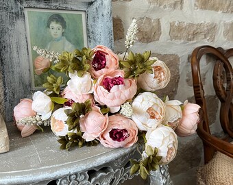 May include: A bouquet of pink and white peonies with dark pink centers rests on a gray, ornate table. A framed portrait of a woman is behind the flowers. The background features a brick wall and a wooden chair.