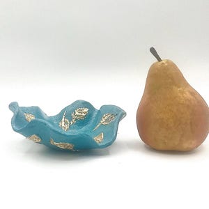 May include: A teal-colored, ruffled bowl with gold leaf accents sits next to a ripe pear. The bowl has a wavy edge and a textured surface. The pear is brown with a reddish blush.