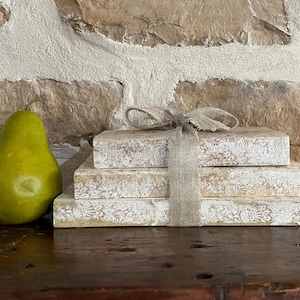 May include: A stack of three rectangular books with distressed white and tan patterned covers, tied together with a light beige fabric ribbon, positioned next to a single smooth green pear on a dark wooden surface.