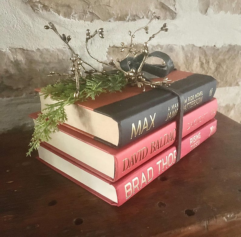 May include: A stack of four books with red and black covers, bound together with a dark leather strap. The top book has the title "MAX" in gold lettering. The stack is decorated with a gold-colored floral accent and greenery.