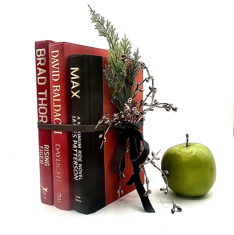 May include: A decorative stack of books bound with a brown leather strap and adorned with greenery and a black ribbon. The books are red and black, with titles including "MAX" and "David Baldacci". A green apple sits nearby.