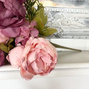 May include: A close-up of a bouquet of artificial flowers, featuring large, soft pink peonies and darker burgundy blooms. Green leaves and stems provide contrast. The flowers are arranged against a white surface with a decorative, ornate border in the background.