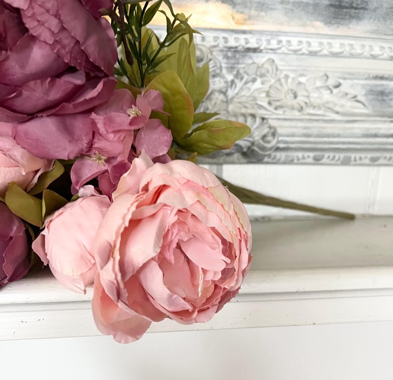 May include: A close-up of a bouquet of artificial flowers, featuring large, soft pink peonies and darker burgundy blooms. Green leaves and stems provide contrast. The flowers are arranged against a white surface with a decorative, ornate border in the background.