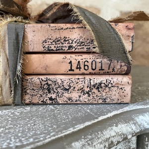 May include: A stack of three rectangular blocks with a light pink finish, wrapped in dark brown frayed ribbon. The top block features cursive script, the middle block displays the black printed number 146017, and the bottom block has a black floral pattern.