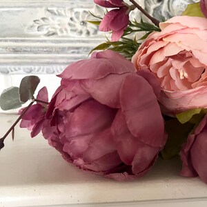 May include: Close-up of artificial flowers, including a large, deep pink peony and a lighter pink peony. Green leaves and stems are visible, with a blurred white background. The flowers are arranged on a white surface.