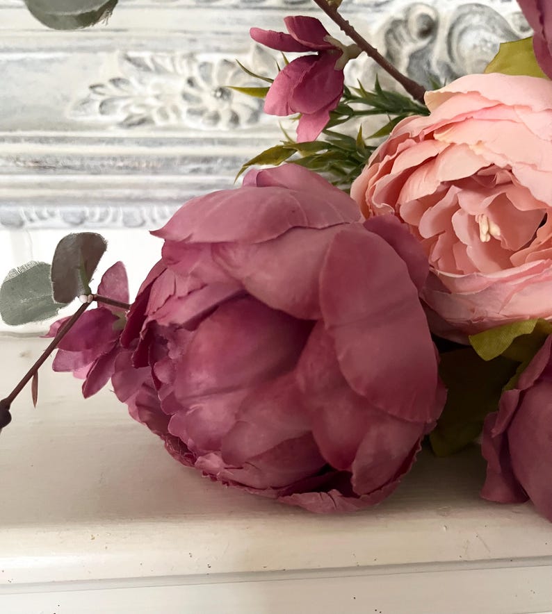 May include: Close-up of artificial flowers, including a large, deep pink peony and a lighter pink peony. Green leaves and stems are visible, with a blurred white background. The flowers are arranged on a white surface.
