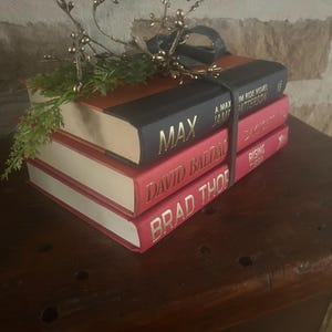 May include: A stack of three hardcover books with red and black covers. The top book is black with the title "MAX" in gold lettering. The other two books are red with gold lettering. A decorative sprig with gold berries and a black ribbon sits on top.