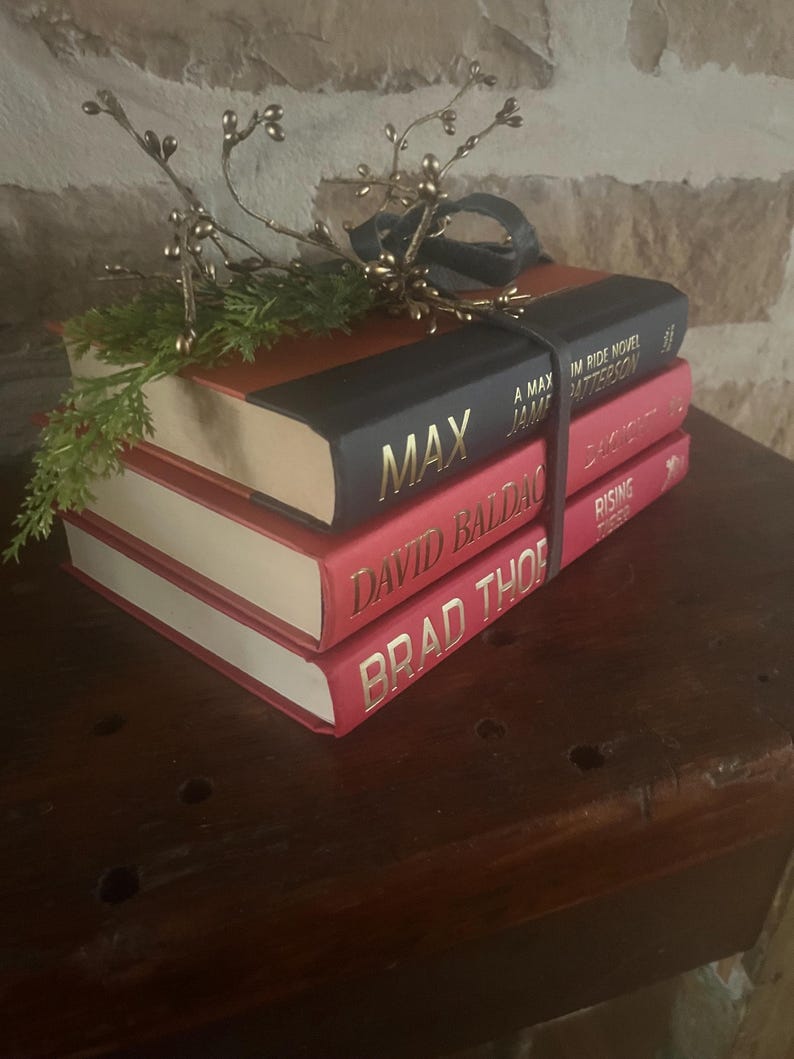 May include: A stack of three hardcover books with red and black covers. The top book is black with the title "MAX" in gold lettering. The other two books are red with gold lettering. A decorative sprig with gold berries and a black ribbon sits on top.