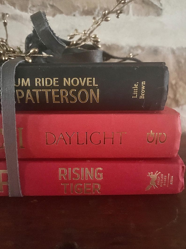 May include: A stack of three books bound together with a gray ribbon. The top book is black with gold lettering that reads "UM RIDE NOVEL PATTERSON." The middle book is red with "DAYLIGHT" in gold. The bottom book is red with "RISING TIGER" in gold.