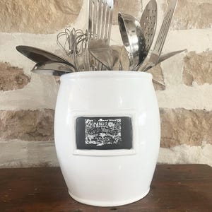 May include: White ceramic utensil holder with a black label that says "Paris Grands Magasins". The holder is filled with various kitchen utensils, including a whisk, a slotted spoon, a spatula, and a ladle.