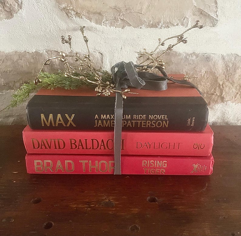 May include: A stack of three books with red and black covers. The top book is black with the title "MAX" and author James Patterson. The other two books are red with gold lettering. A gray ribbon and decorative sprigs are on top.