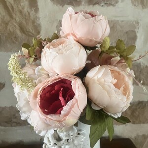 May include: A bouquet of pink and red peonies with green leaves in a white vase. The peonies are arranged in a cluster, with the pink peonies on top and the red peony in the center.
